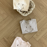 Drei gefaltete Bebe Holic Smile Tees aus weicher Baumwolle mit Smileys darauf sind auf einem Holzboden angeordnet. Zwei T-Shirts liegen direkt auf dem Boden, während eines in einem geflochtenen Korb liegt. Die T-Shirts sind in hellen Creme-, Grau- und Rosatönen gehalten.