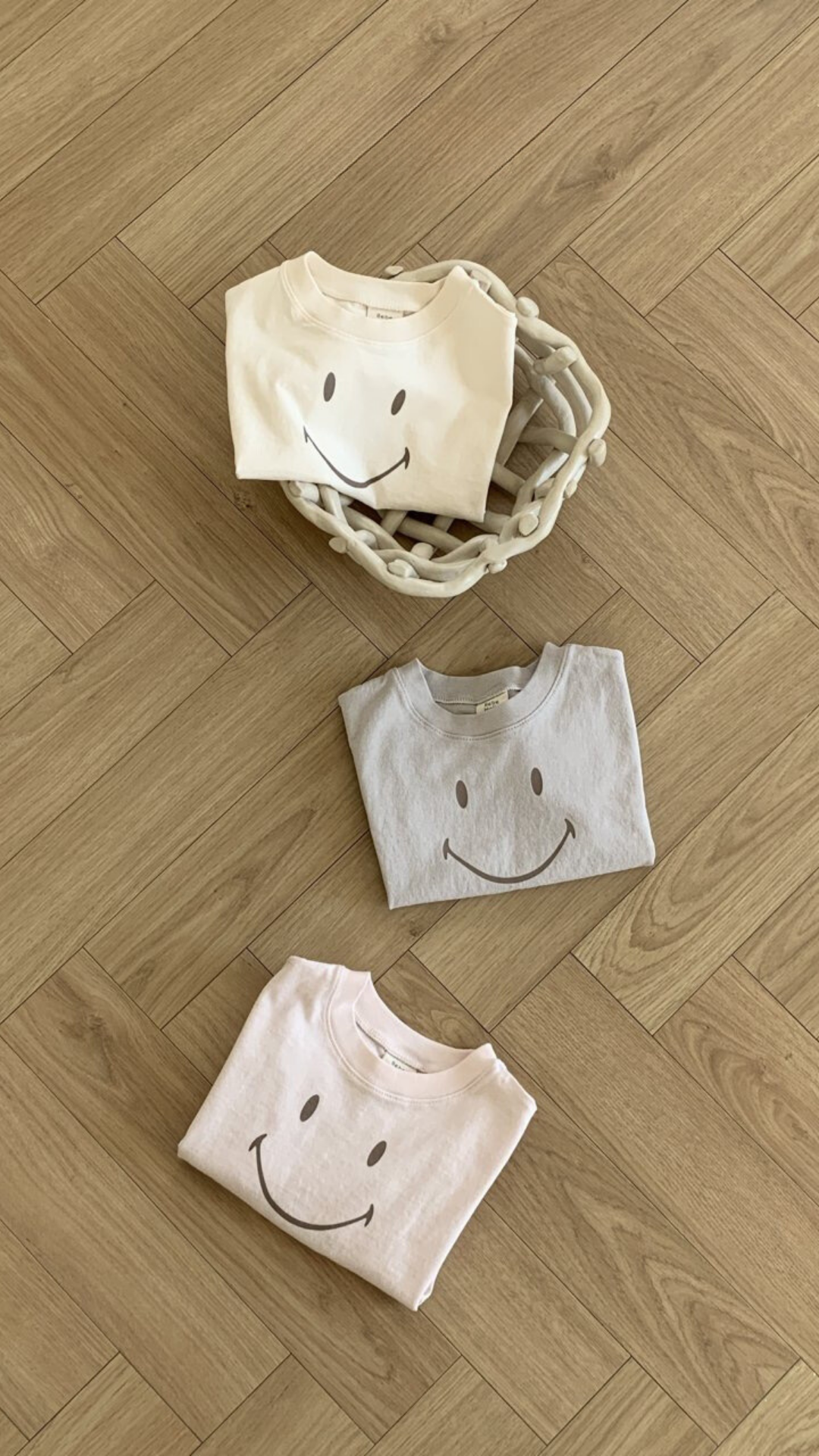 Drei gefaltete Bebe Holic Smile Tees aus weicher Baumwolle mit Smileys darauf sind auf einem Holzboden angeordnet. Zwei T-Shirts liegen direkt auf dem Boden, während eines in einem geflochtenen Korb liegt. Die T-Shirts sind in hellen Creme-, Grau- und Rosatönen gehalten.