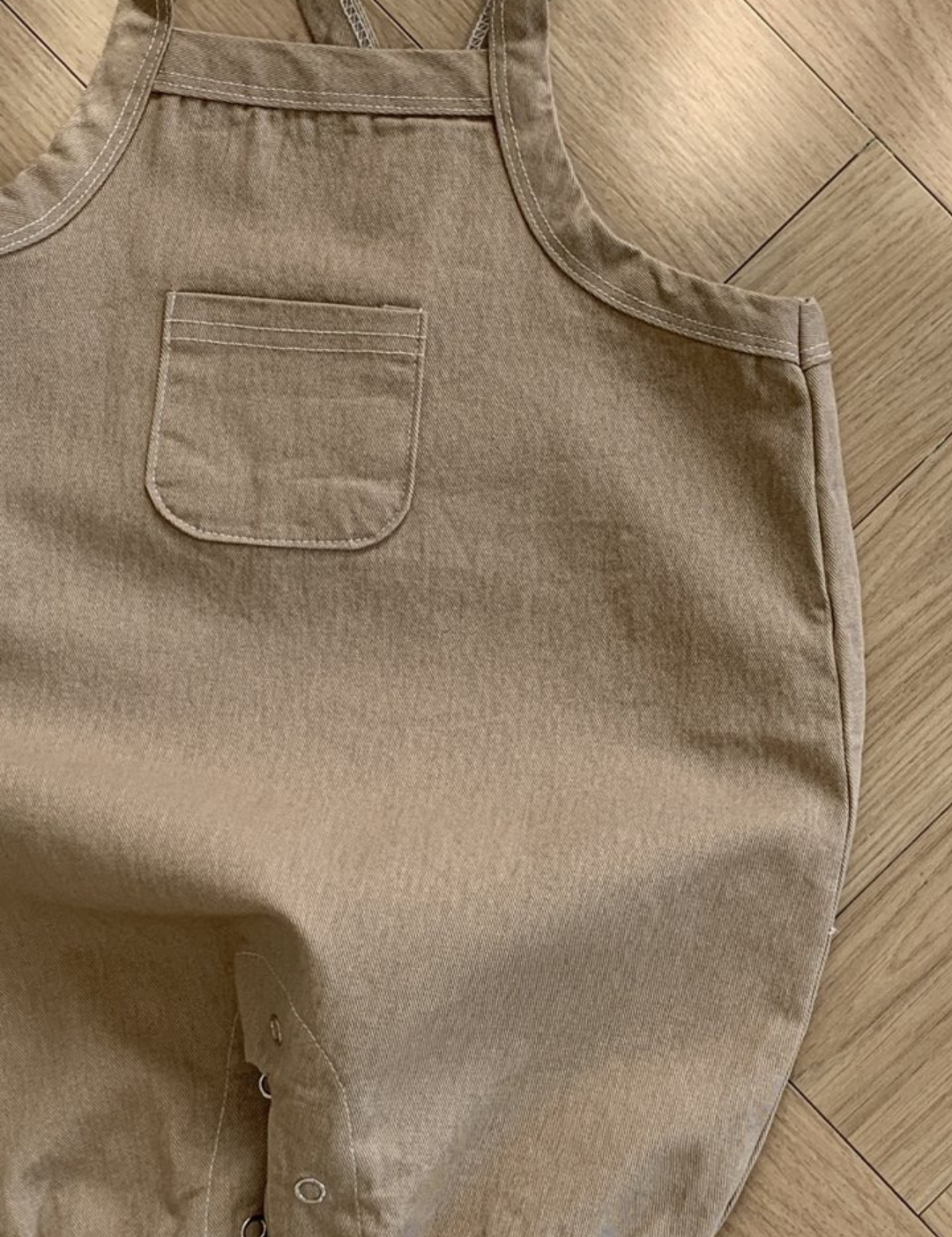 Ein hellbrauner Bebe Holic Twill-Body mit verstellbarem Träger und einer kleinen Brusttasche. Der Strampler aus weicher Baumwolle hat Druckknöpfe an der Unterseite zum einfachen Windelwechseln. Er liegt flach auf einem Holzboden, wobei Licht und Schatten auf der Oberfläche sichtbar sind.