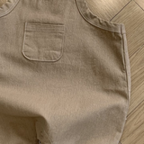 Ein hellbrauner Bebe Holic Twill-Body mit verstellbarem Träger und einer kleinen Brusttasche. Der Strampler aus weicher Baumwolle hat Druckknöpfe an der Unterseite zum einfachen Windelwechseln. Er liegt flach auf einem Holzboden, wobei Licht und Schatten auf der Oberfläche sichtbar sind.