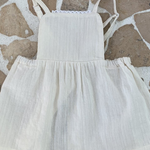 Ein ärmelloses, cremefarbenes Kleinkinderkleid von Bebe Holic mit quadratischem Ausschnitt und Spitzenbesatz. Das Kleid hat dünne Träger und einen gerafften, ausgestellten Lagenrock. Es liegt flach auf einer gepflasterten Oberfläche, wobei weiße Fugenlinien unregelmäßige Muster bilden und seine eleganten Details zur Geltung bringen.