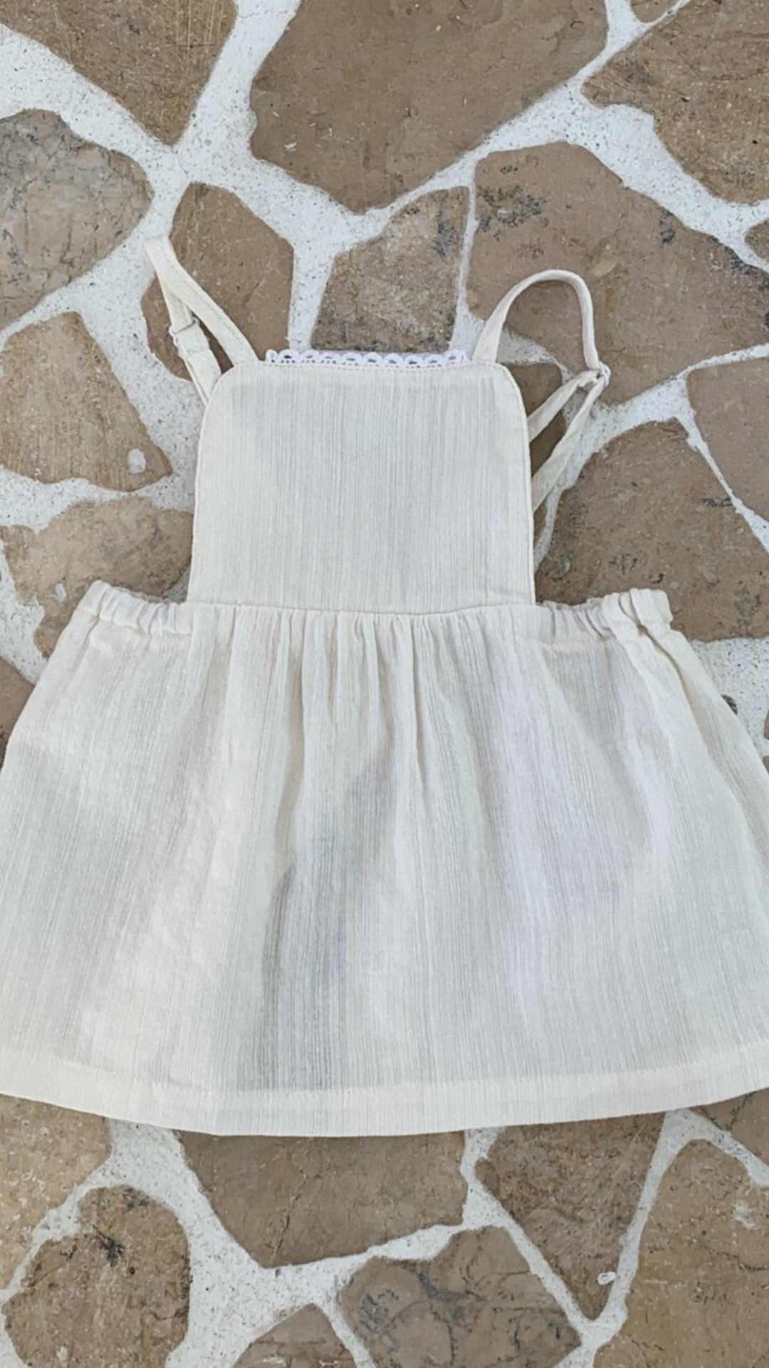 Ein ärmelloses, cremefarbenes Kleinkinderkleid von Bebe Holic mit quadratischem Ausschnitt und Spitzenbesatz. Das Kleid hat dünne Träger und einen gerafften, ausgestellten Lagenrock. Es liegt flach auf einer gepflasterten Oberfläche, wobei weiße Fugenlinien unregelmäßige Muster bilden und seine eleganten Details zur Geltung bringen.