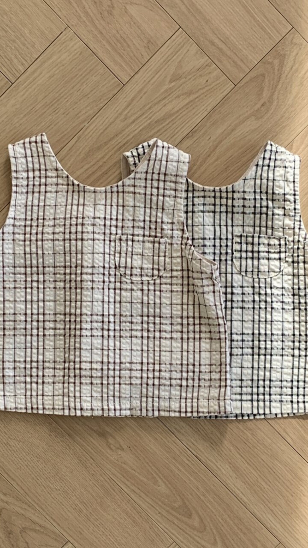 Zwei Summer Check Sleeveless Tees von Bebe Holic aus strukturiertem Stoff mit Gittermuster liegen auf einem Holzboden. Ein Oberteil ist weiß mit rotem Gitter, das andere weiß mit schwarzem Gitter. Jedes Oberteil hat eine kleine Tasche auf der Brust, die einen Hauch modischen Flairs verleiht und perfekt in jede Bebeholic-Kollektion passt.