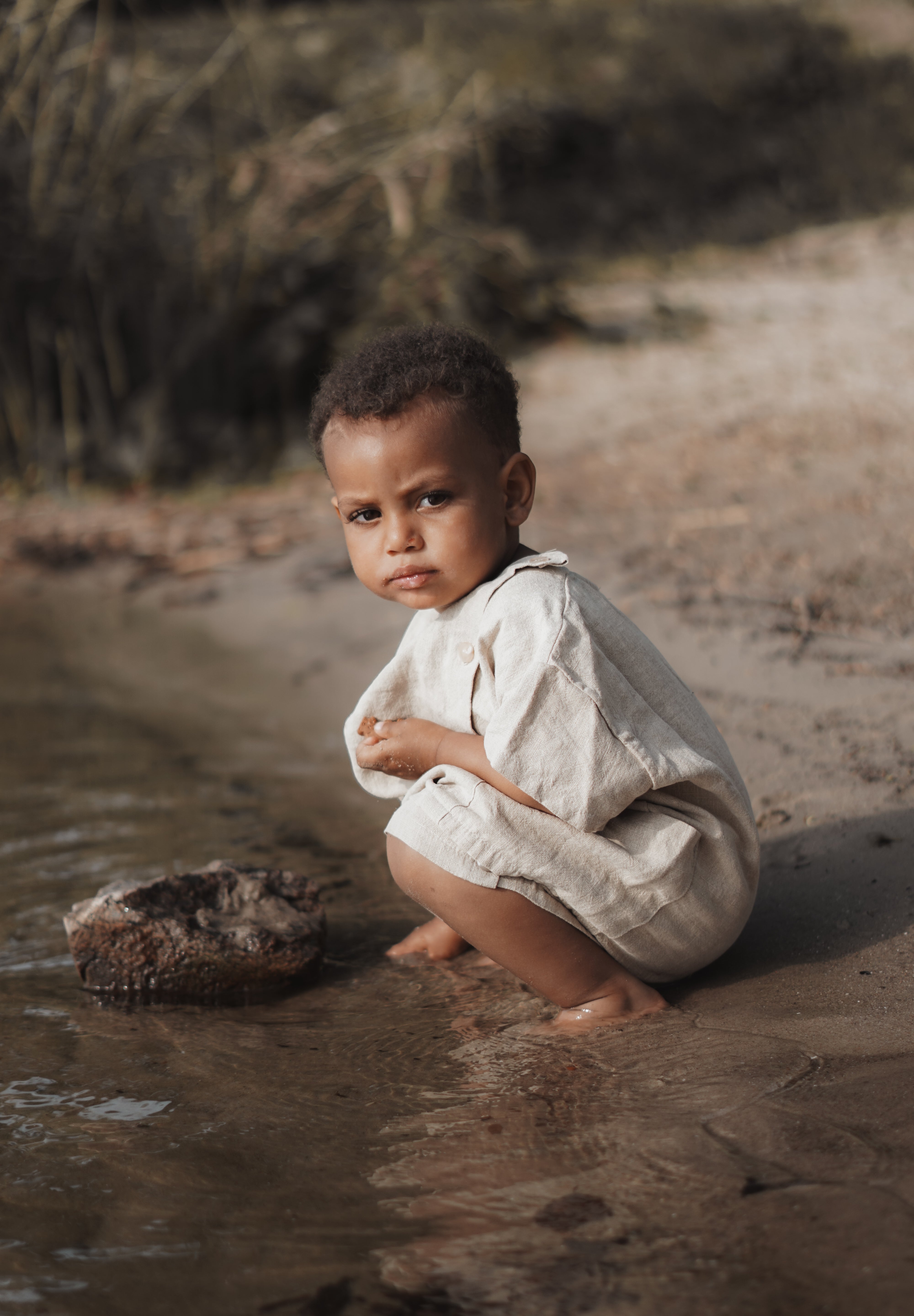 Ein kleines Kind in einem hellen Cinamon Top Bottom Set von Anggo hockt am Rand eines Gewässers und blickt aufmerksam in die Kamera. Der Hintergrund ist eine natürliche Umgebung im Freien mit Sandboden, Wasser und Vegetation.