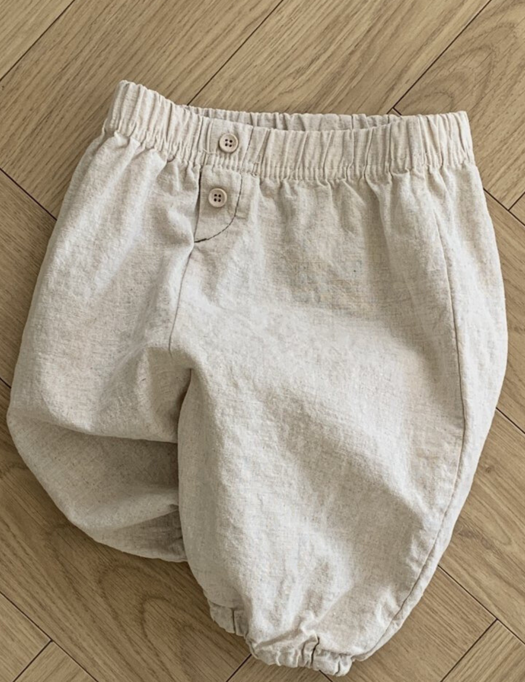 Ein Paar beige Bebe Holic Natural Pants mit elastischer Taille und zwei Knöpfen auf der Vorderseite, flach auf einem hellen Holzboden mit Fischgrätenmuster. Diese vielseitigen Must-have-Shorts sind ein Muss für die Garderobe Ihres Babys.