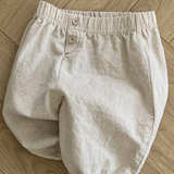 Ein Paar beige Bebe Holic Natural Pants mit elastischer Taille und zwei Knöpfen auf der Vorderseite, flach auf einem hellen Holzboden mit Fischgrätenmuster. Diese vielseitigen Must-have-Shorts sind ein Muss für die Garderobe Ihres Babys.