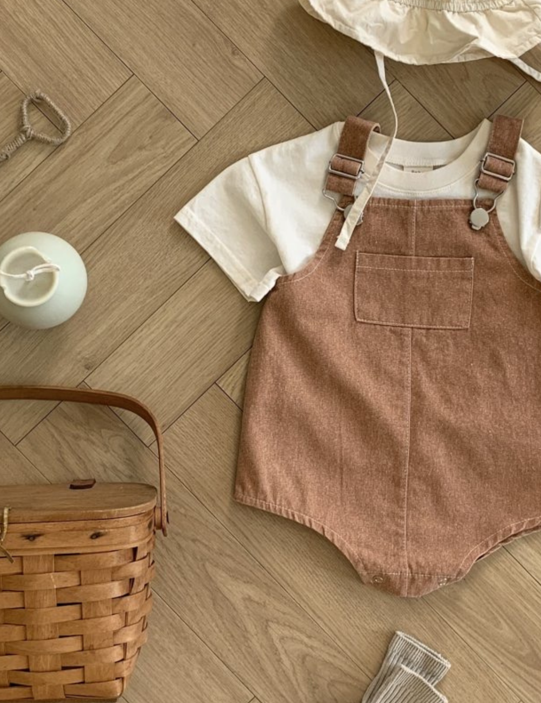 Ein bereitgelegtes Outfit für ein Kind, darunter ein weißes Kurzarmhemd, ein brauner Bebe Holic Pigment Latzhosen-Body mit verstellbaren Trägern, ein weißer Sonnenhut und eine einzelne beige Socke. Um das Outfit herum stehen ein Weidenkorb, eine Spielzeugrassel und eine kleine weiße Vase, alles auf einem Holzboden.