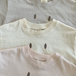 Drei gefaltete Bebe Holic Smile Tees aus weicher Baumwolle liegen gestapelt auf einer Holzfläche. Von oben nach unten sind die Farben hellgrau, cremefarben und hellrosa.