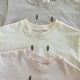 Drei gefaltete Bebe Holic Smile Tees aus weicher Baumwolle liegen gestapelt auf einer Holzfläche. Von oben nach unten sind die Farben hellgrau, cremefarben und hellrosa.