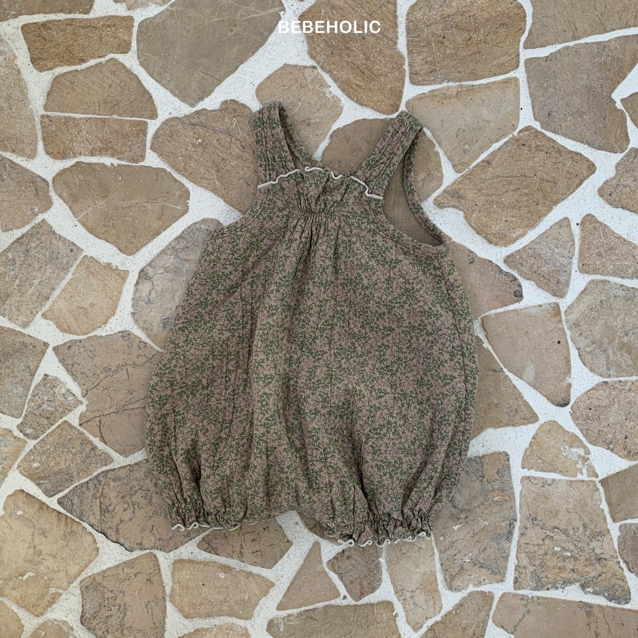 Ein Karen-Bodysuit mit Blumenmuster, elastischen Beinöffnungen, gerüschten Rändern und Schulterriemen liegt flach auf einer Steinfliesenoberfläche. Der aus atmungsaktiver Baumwolle gefertigte Stoff zeigt dicht gepackte, winzige rosa Blumen auf einem gedämpften grünen Hintergrund. Über dem Anzug steht „Bebe Holic“.