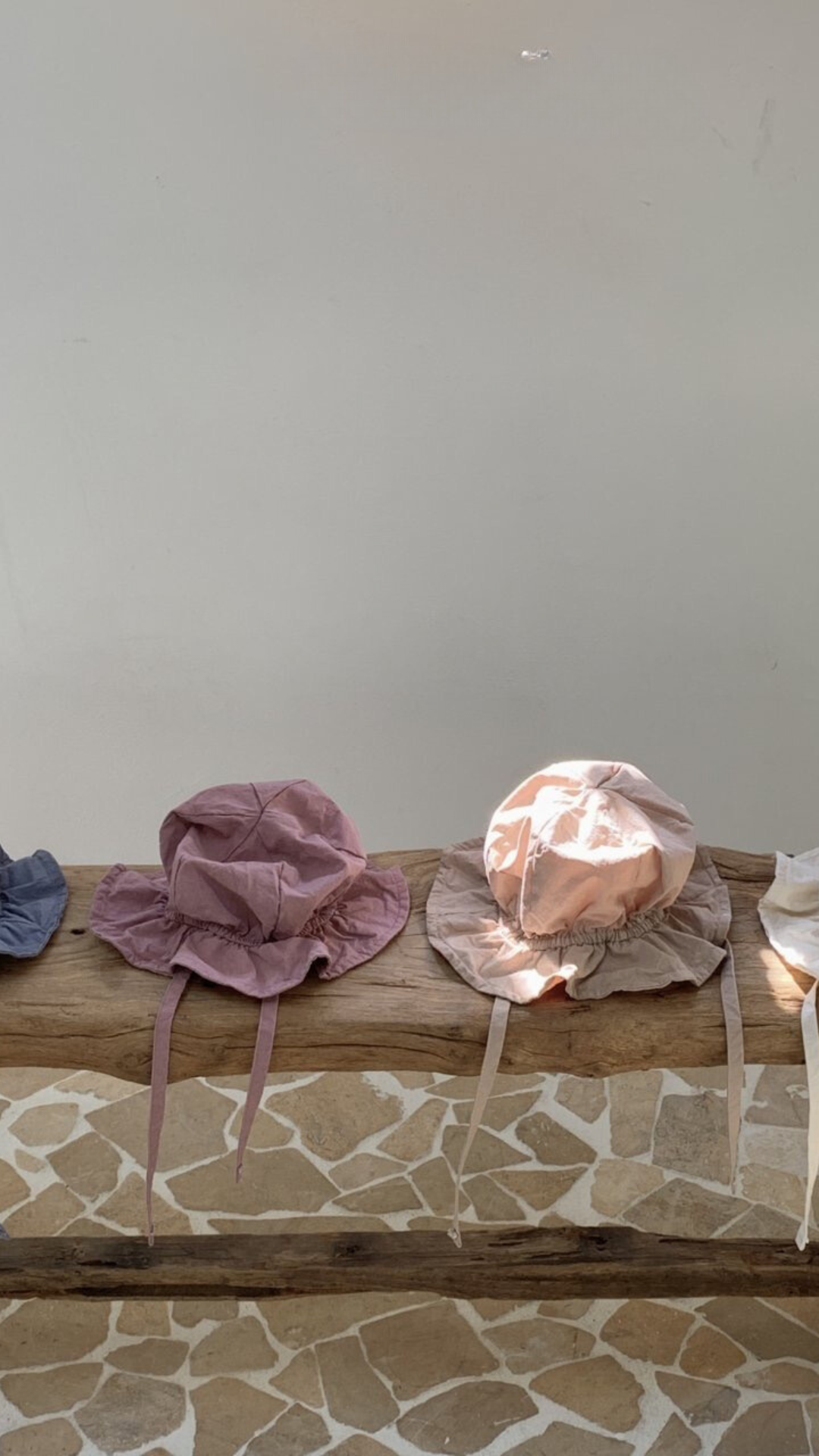 Eine Reihe pastellfarbener Crunch Bucket Hats mit breiter Krempe und Kinnriemen wird auf einem Holzstamm vor einem Hintergrund mit strukturiertem Steinmuster und einer schlichten weißen Wand präsentiert. Die Bebe Holic-Hüte in den Farbtönen Lavendel, Hellrosa und Creme zeigen eine natürliche Farbpalette.