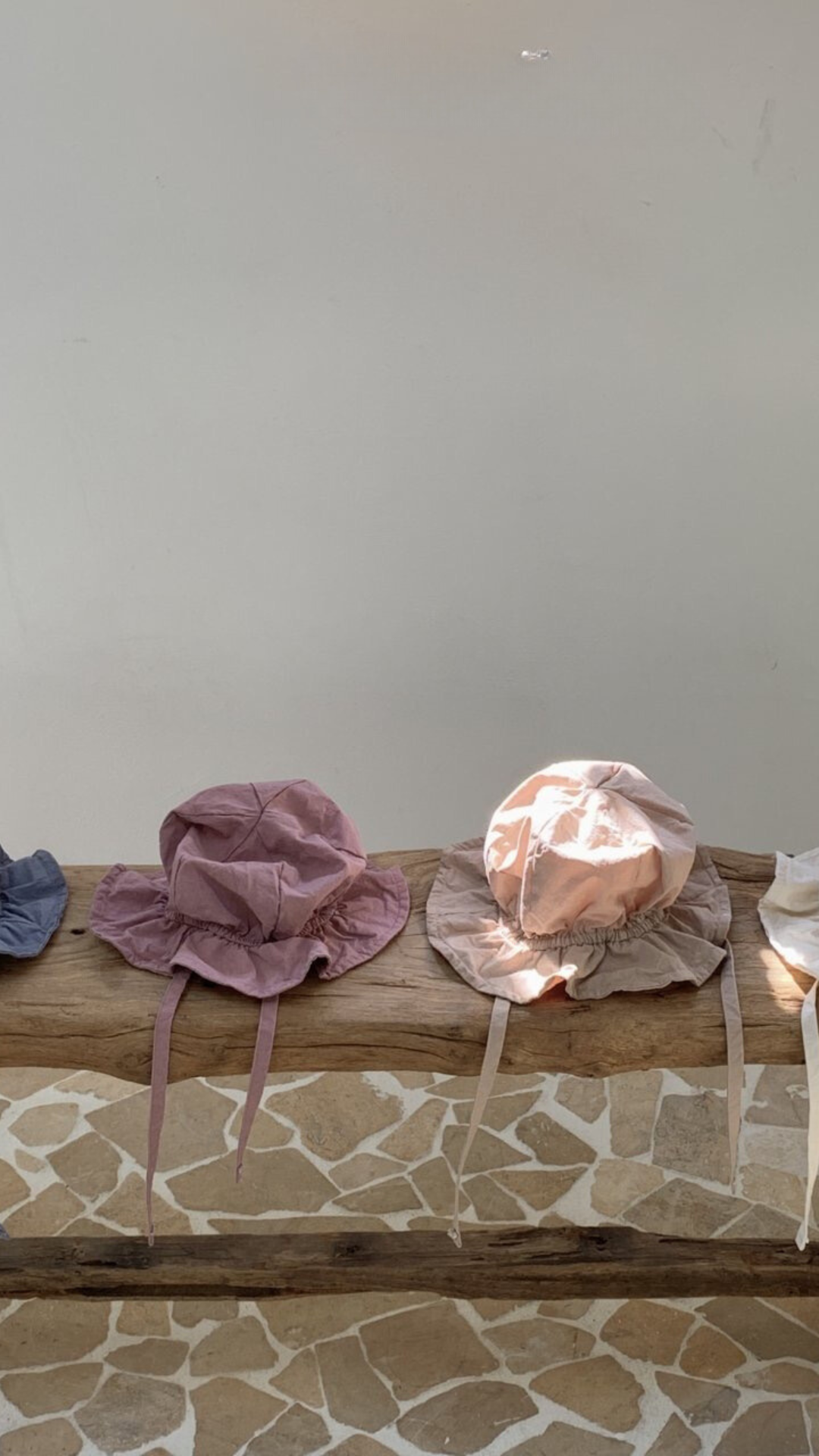 Zwei Crunch Bucket Hats von Bebe Holic mit Kinnriemen werden auf einer Holzoberfläche präsentiert. Der Hut links ist malvenfarben und der rechte hellpfirsichfarben. Der Hintergrund besteht aus einer hellgrauen Wand und beige-braunen Steinfliesen und bietet eine natürliche Farbpalette, die perfekt zu jedem Anlass von Bebe Holic passt.