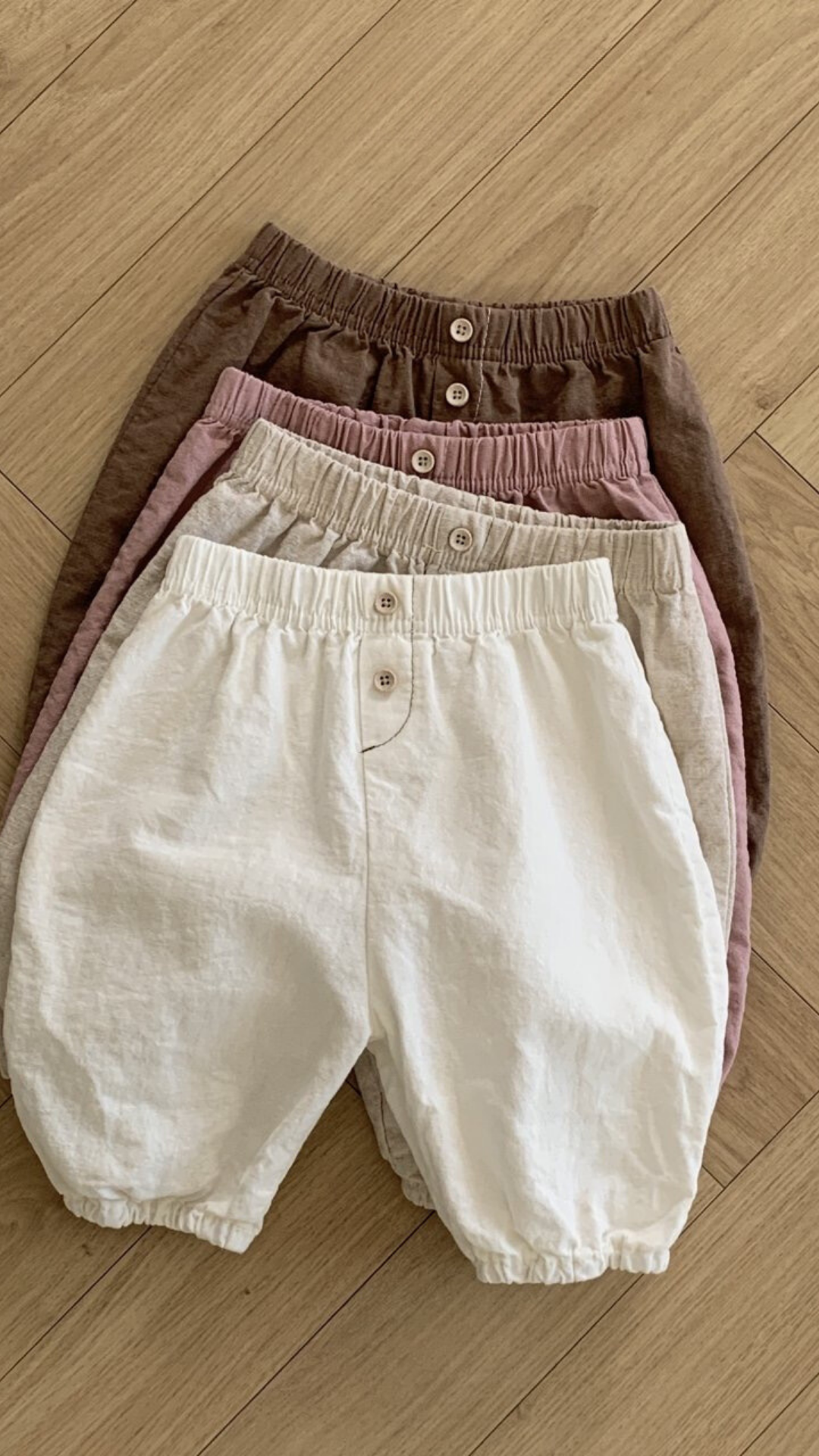 Vier Paar Natural Pants für Kinder von Bebe Holic liegen ordentlich gestapelt auf einem Holzboden. Diese vielseitigen Must-have-Shorts sind in den Farben Braun, Rosa, Beige und Weiß erhältlich und verfügen jeweils über eine elastische Taille und einen Knopf in der Mitte vorne.