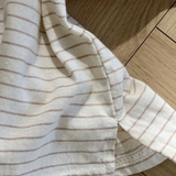 Eine Nahaufnahme eines weißen Stoffes mit dünnen, hellbraunen Streifen auf einer Holzoberfläche. Der Stoff scheint Teil eines Kleidungsstückes zu sein, möglicherweise das Basic ST Tee von Bebe Holic, mit sichtbarer Naht und teilweisem Ärmel.