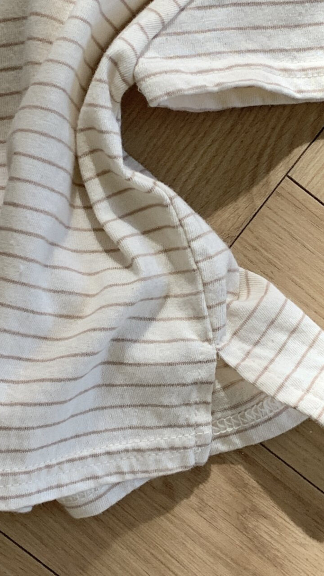 Eine Nahaufnahme eines weißen Stoffes mit dünnen, hellbraunen Streifen auf einer Holzoberfläche. Der Stoff scheint Teil eines Kleidungsstückes zu sein, möglicherweise das Basic ST Tee von Bebe Holic, mit sichtbarer Naht und teilweisem Ärmel.