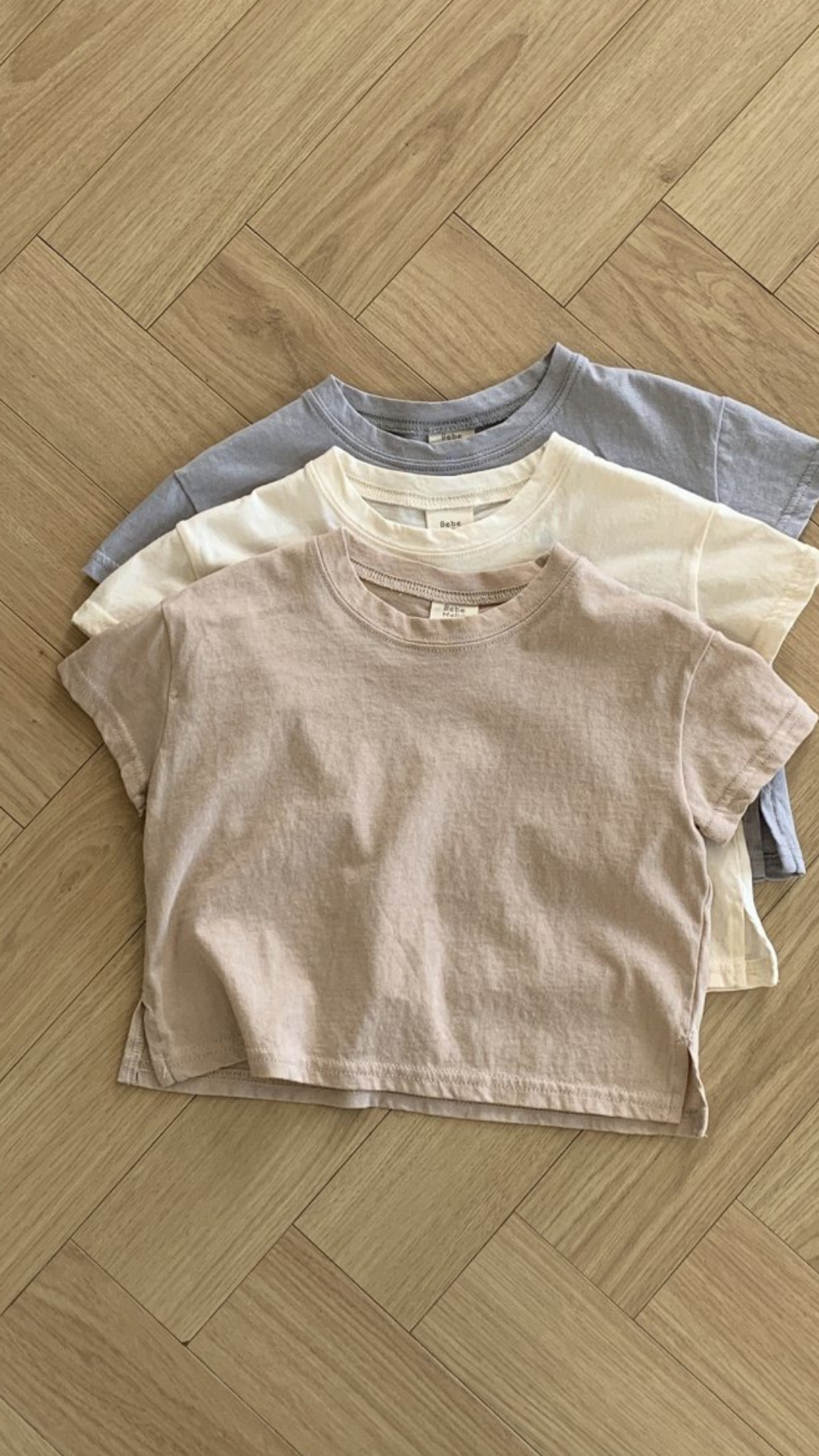 Drei pastellfarbene Basic-T-Shirts von Bebe Holic in Beige-, Creme- und Hellgrautönen sind ordentlich auf einem hölzernen Boden mit Fischgrätenmuster gestapelt. Diese T-Shirts aus hochwertiger Baumwolle haben eine lässige, entspannte Passform und kurze Ärmel in natürlichen Tönen.