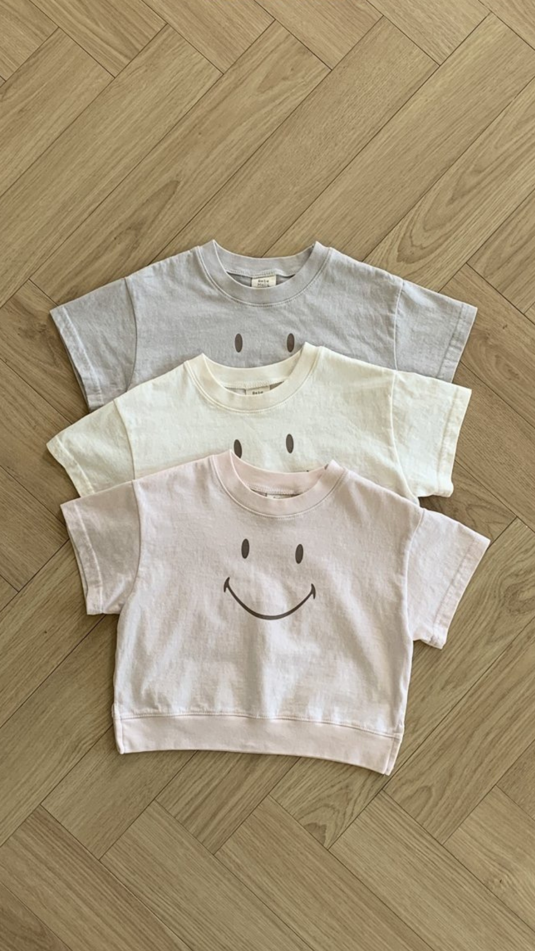 Drei kurzärmlige Bebe Holic Smile T-Shirts liegen ordentlich gestapelt auf einem Holzboden. Das obere Shirt ist hellgrau, das mittlere ist cremefarben und das untere ist hellrosa. Jedes dieser Kinder-T-Shirts aus weicher Baumwolle hat auf der Vorderseite ein einfaches Smiley-Design.