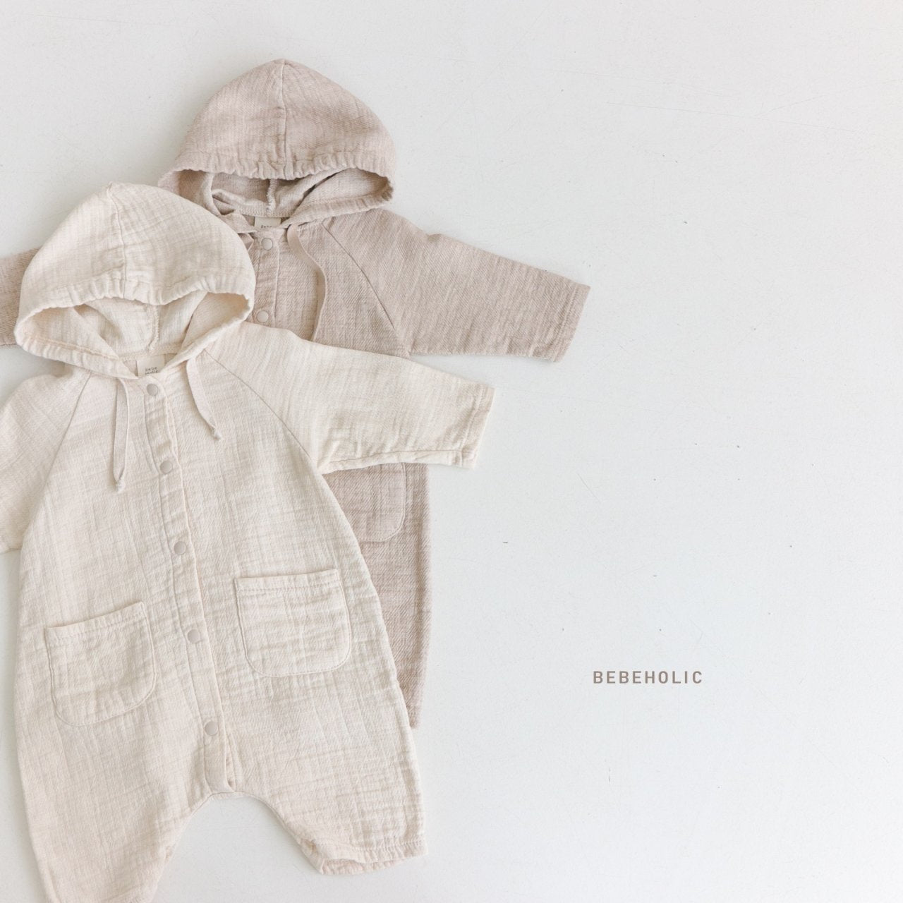 Zwei Baby-Kapuzenstrampler, oder wie wir sie nennen: Hoody-Bodys, sind auf weißem Hintergrund ausgelegt. Ein Strampler ist cremefarben und der andere hellbeige. Beide Strampler haben Vordertaschen, lange Ärmel und Knopfverschlüsse. Der Markenname „Bebe Holic“ ist unter den Stramplern aufgedruckt und sorgt für Babykomfort für Ihren Kleinen.