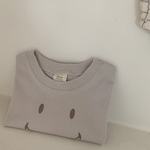 Ein ordentlich gefaltetes hellgraues T-Shirt aus weicher Baumwolle mit einem einfachen, braunen Smiley-Design auf der Vorderseite liegt auf einem weißen Regal vor einer hellen Wand. Auf der rechten Seite des Bildes hängt ein kleines Stück gemusterter Stoff, auf dem das Bebe Holic Smile Tee zu sehen ist.