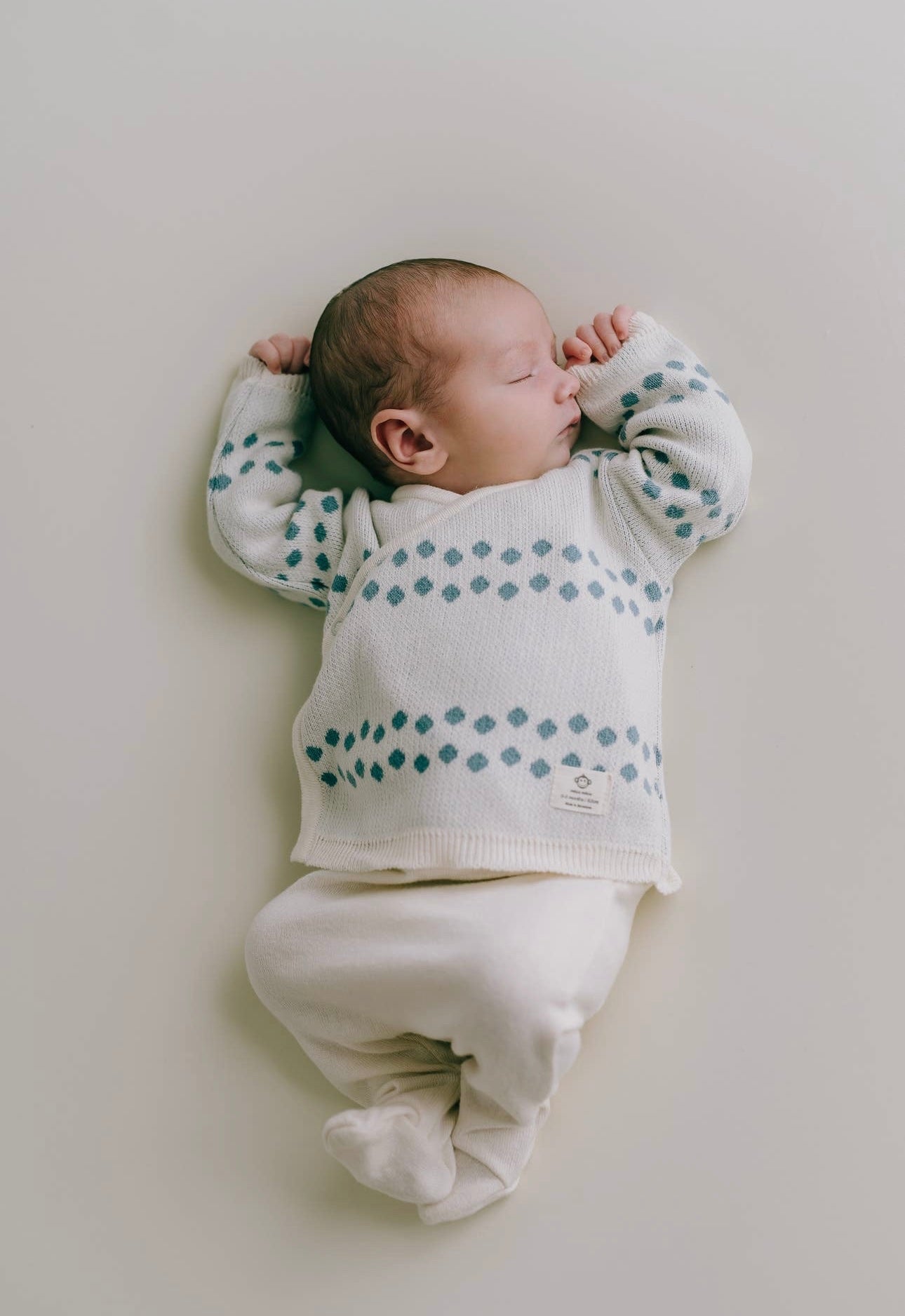 Ein Neugeborenes schläft friedlich auf einem hellen Hintergrund. Es trägt einen weißen Pullover mit blauen Punkten und dazu passende Hosen von Pack Dots Jeans für Neugeborene, alles aus 100 % Bio-Baumwolle. Das Baby hat seine Hände in der Nähe seines Micu Micu ausgestreckt.