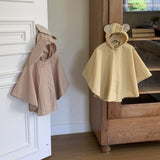 Zwei Bebe Holic Tierregenmäntel 6m-24m - Der perfekte Regenponcho für kleine Abenteurer hängen nebeneinander. Der linke Poncho ist beige mit Hasenohren an der Kapuze und der rechte Poncho ist gelb mit Bärenohren an der Kapuze. Sie hängen vor einer weißen Tür und einem Holzschrank und zeigen ihre bequeme Passform in einem gemütlichen Zimmer im Hintergrund.