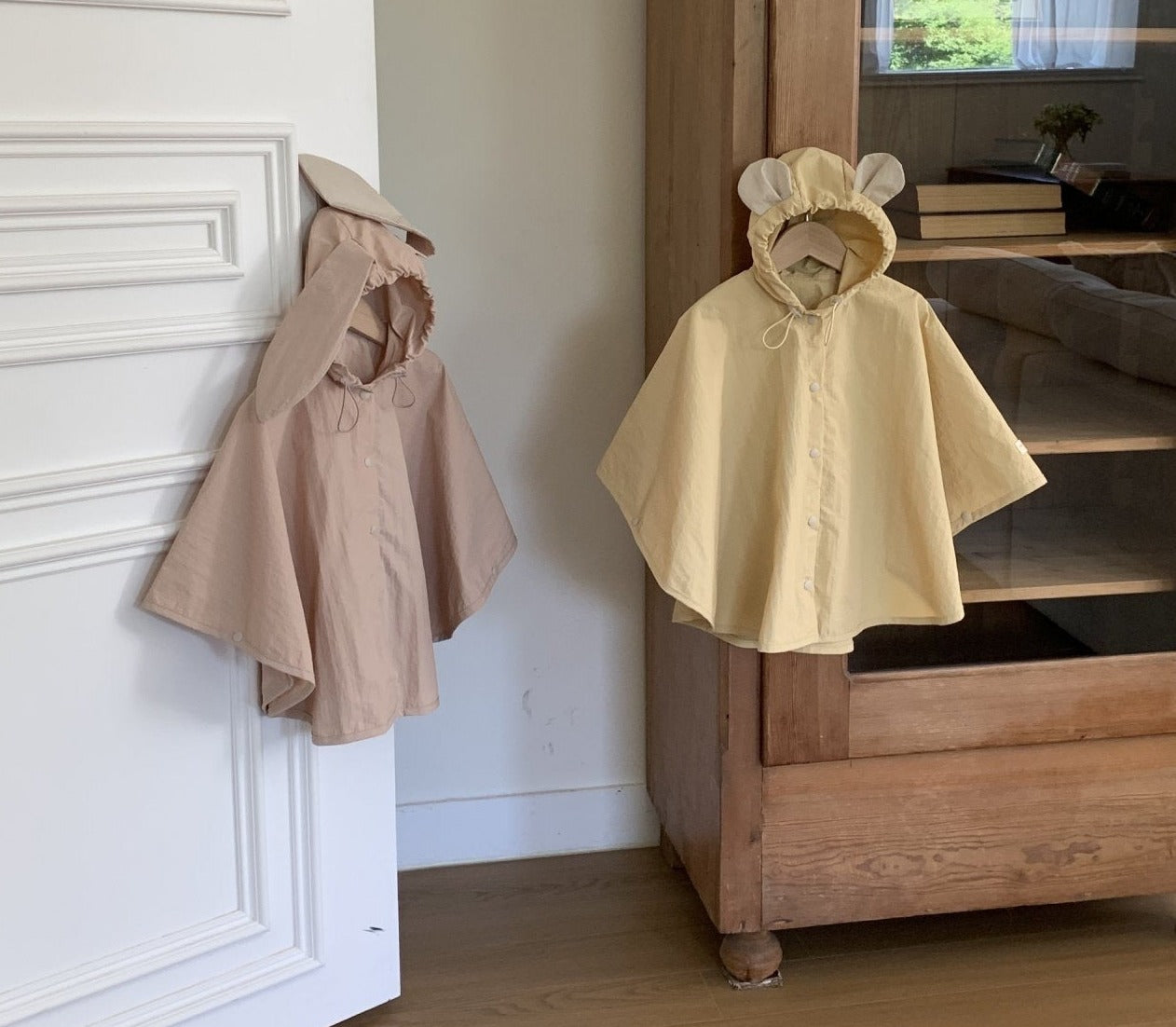 Zwei Bebe Holic Tierregenmäntel 6m-24m - Der perfekte Regenponcho für kleine Abenteurer hängen nebeneinander. Der linke Poncho ist beige mit Hasenohren an der Kapuze und der rechte Poncho ist gelb mit Bärenohren an der Kapuze. Sie hängen vor einer weißen Tür und einem Holzschrank und zeigen ihre bequeme Passform in einem gemütlichen Zimmer im Hintergrund.