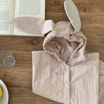 Ein rosa **Tierregenmantel 6m-24m - Der perfekte Regenponcho für kleine Abenteurer** mit Hasenohrenkapuze liegt zusammengefaltet auf einem Holztisch neben einem offenen Buch. Ein durchsichtiges Trinkglas und ein Teller mit ein paar gelben Früchten stehen ebenfalls auf dem Tisch. Der Text „BEBEHOLIC“ ist unten im Bild zu sehen und unterstreicht seine bequeme Passform.