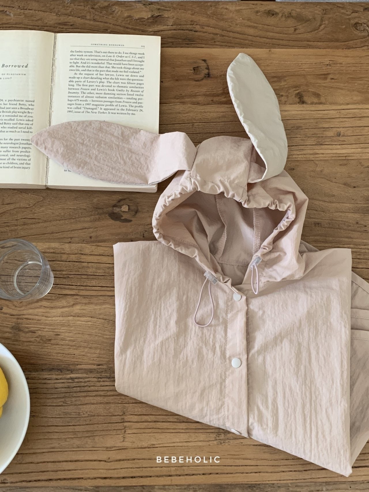 Ein rosa **Tierregenmantel 6m-24m - Der perfekte Regenponcho für kleine Abenteurer** mit Hasenohrenkapuze liegt zusammengefaltet auf einem Holztisch neben einem offenen Buch. Ein durchsichtiges Trinkglas und ein Teller mit ein paar gelben Früchten stehen ebenfalls auf dem Tisch. Der Text „BEBEHOLIC“ ist unten im Bild zu sehen und unterstreicht seine bequeme Passform.