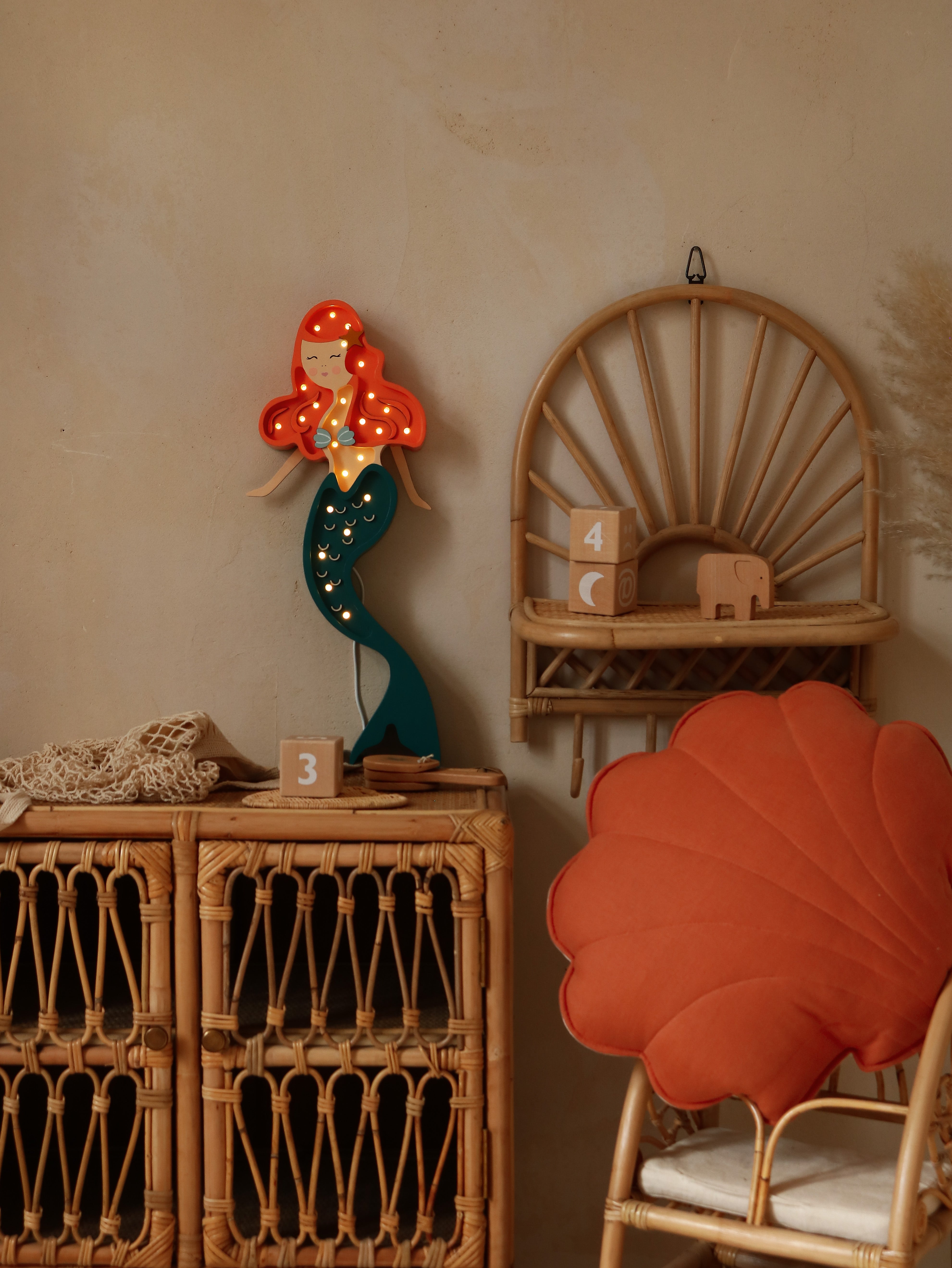 Eine gemütliche Zimmerecke mit einer Little Lights Ariel Mermaid-Lampe auf einem Holzhocker, einem Bambusstuhl mit einem korallenblattförmigen Kissen und einem Rattanschrank mit den Schubladen 3 und 4.