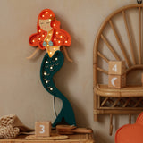 Eine Little Lights Ariel Mermaid Lampe mit roten Haaren und blaugrünem Schwanz ist an einer beigen Wand montiert, umgeben von Korbmöbeln und Dekorationsobjekten.