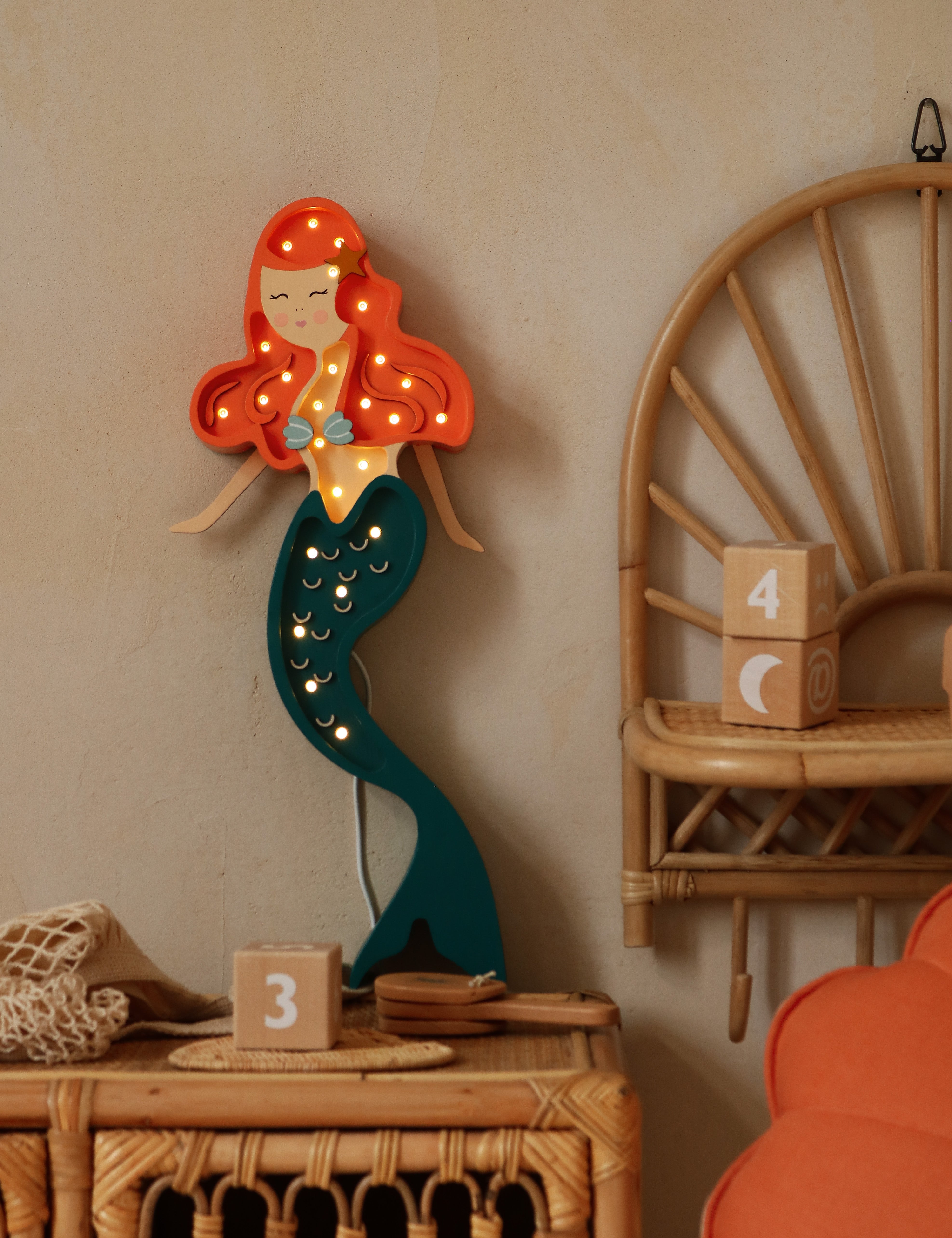 Eine Little Lights Ariel Mermaid Lampe mit roten Haaren und blaugrünem Schwanz ist an einer beigen Wand montiert, umgeben von Korbmöbeln und Dekorationsobjekten.