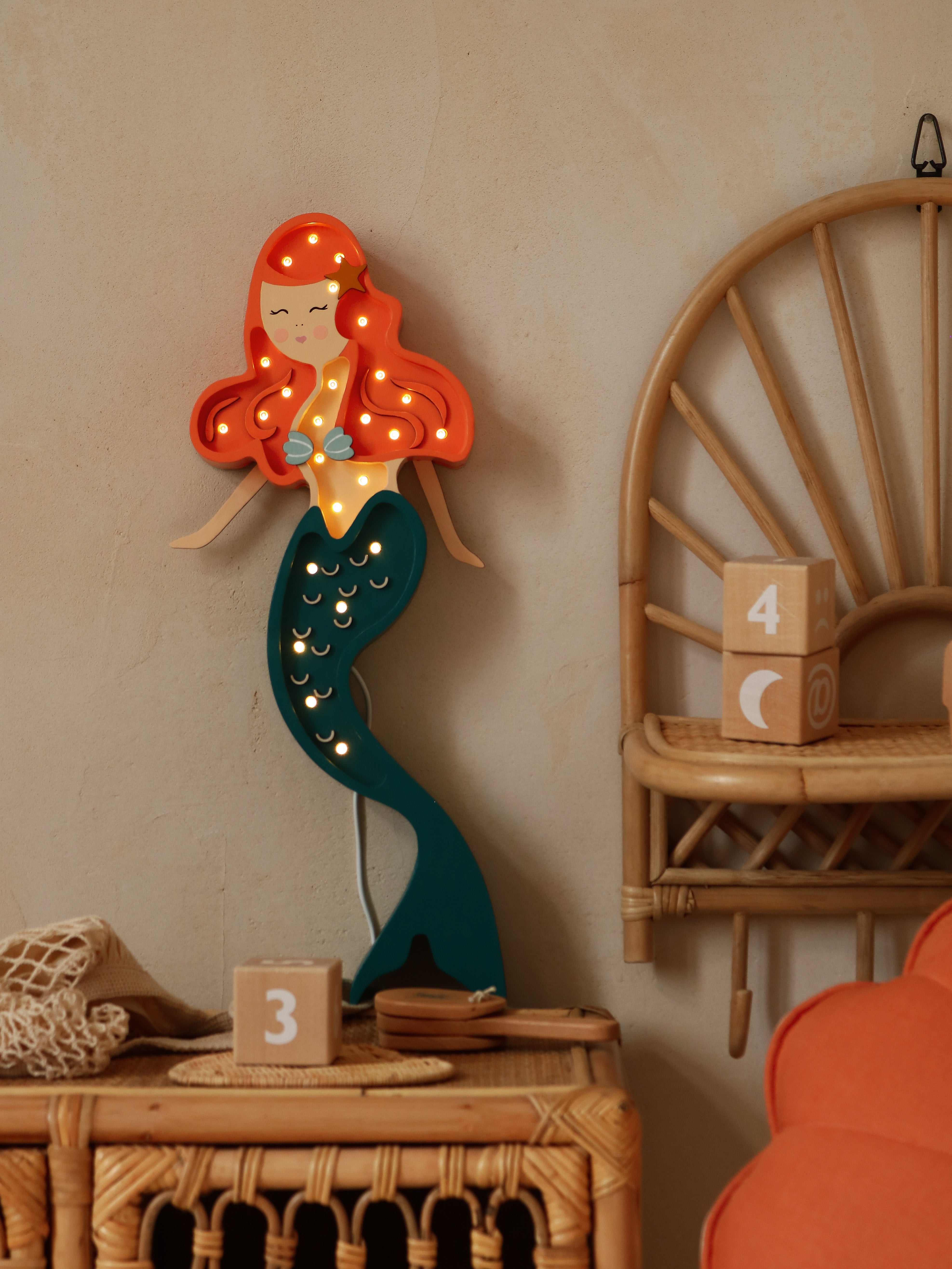 Eine Little Lights Ariel Mermaid Lampe mit roten Haaren und blaugrünem Schwanz ist an einer beigen Wand montiert, umgeben von Korbmöbeln und Dekorationsobjekten.