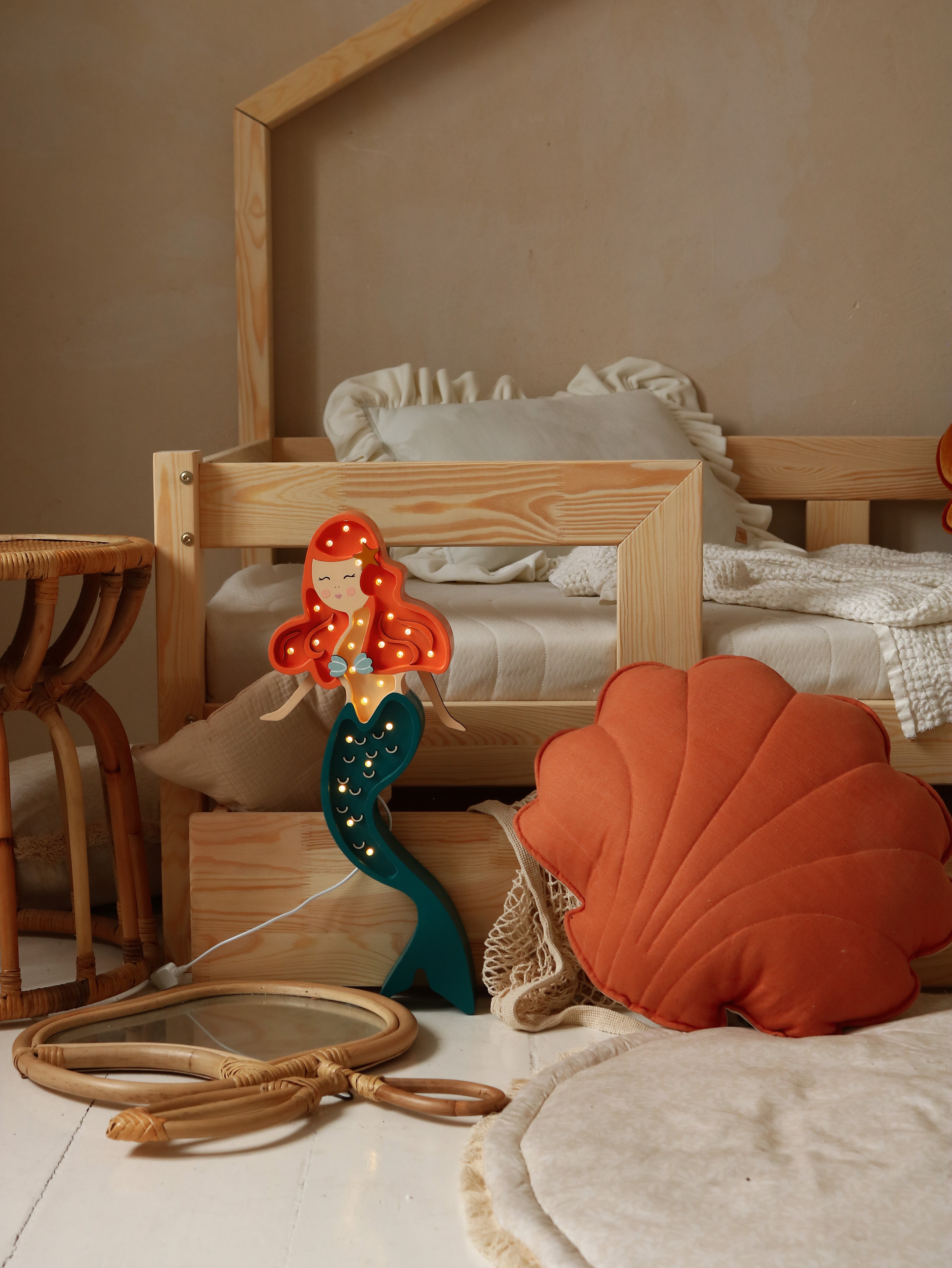 Eine gemütliche Kinderzimmerecke mit einem Etagenbett aus Holz mit weißer Bettwäsche, einer Ariel-Meerjungfrau-Lampe von Little Lights, einem orangefarbenen Kissen in Muschelform und einem ausgelegten Seil.