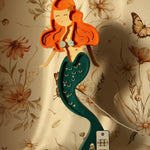 Eine skurrile Illustration einer Meerjungfrau mit orangefarbenem Haar und einem grün gefleckten Schwanz, die eine Fernbedienung hält, positioniert vor einem Hintergrund mit Blumen- und Schmetterlingsmuster mit der Little Lights Ariel Mermaid Lampe – Zauberhaftes Nachtlicht für Kinderzimmer.