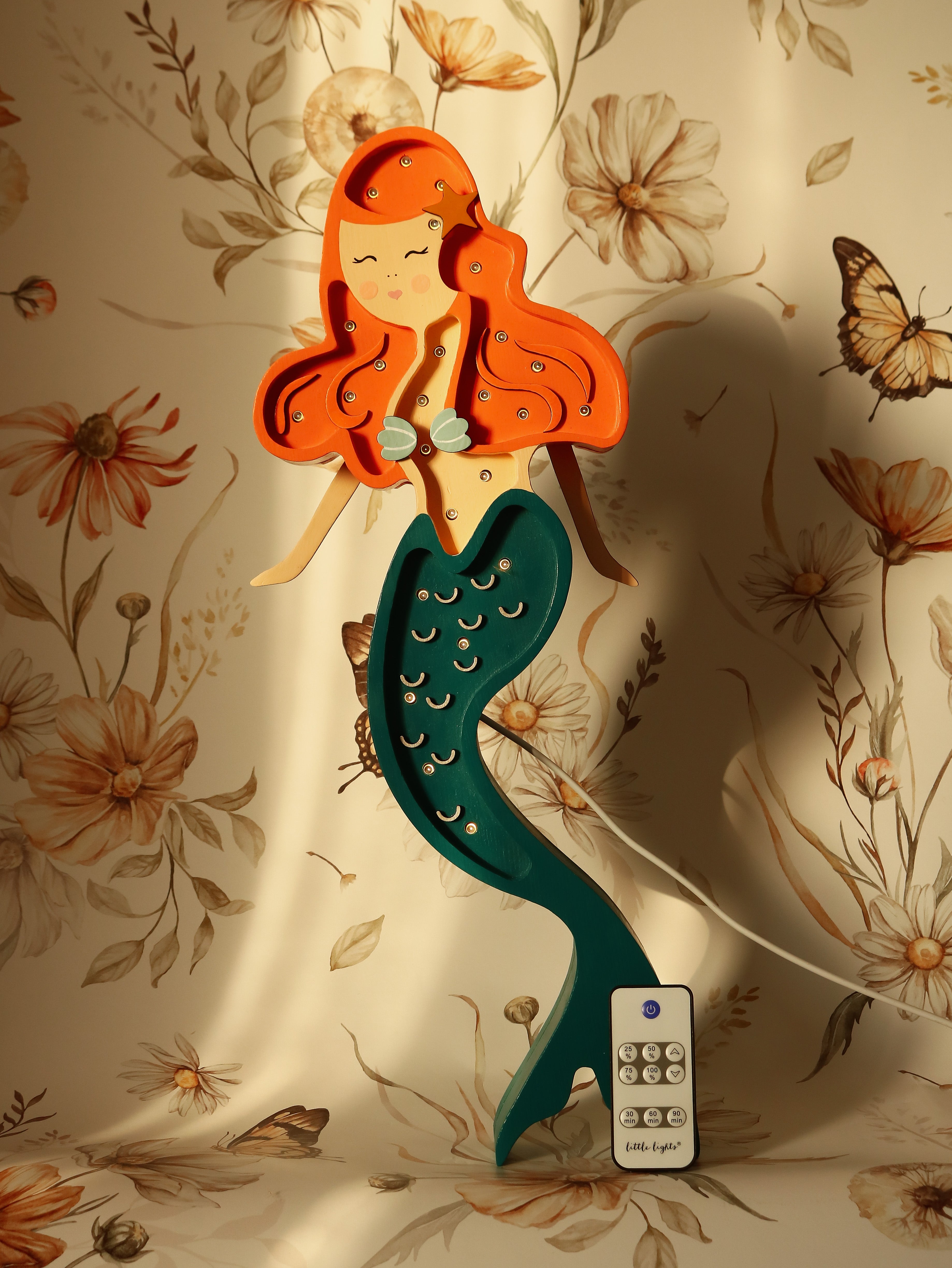 Eine skurrile Illustration einer Meerjungfrau mit orangefarbenem Haar und einem grün gefleckten Schwanz, die eine Fernbedienung hält, positioniert vor einem Hintergrund mit Blumen- und Schmetterlingsmuster mit der Little Lights Ariel Mermaid Lampe – Zauberhaftes Nachtlicht für Kinderzimmer.