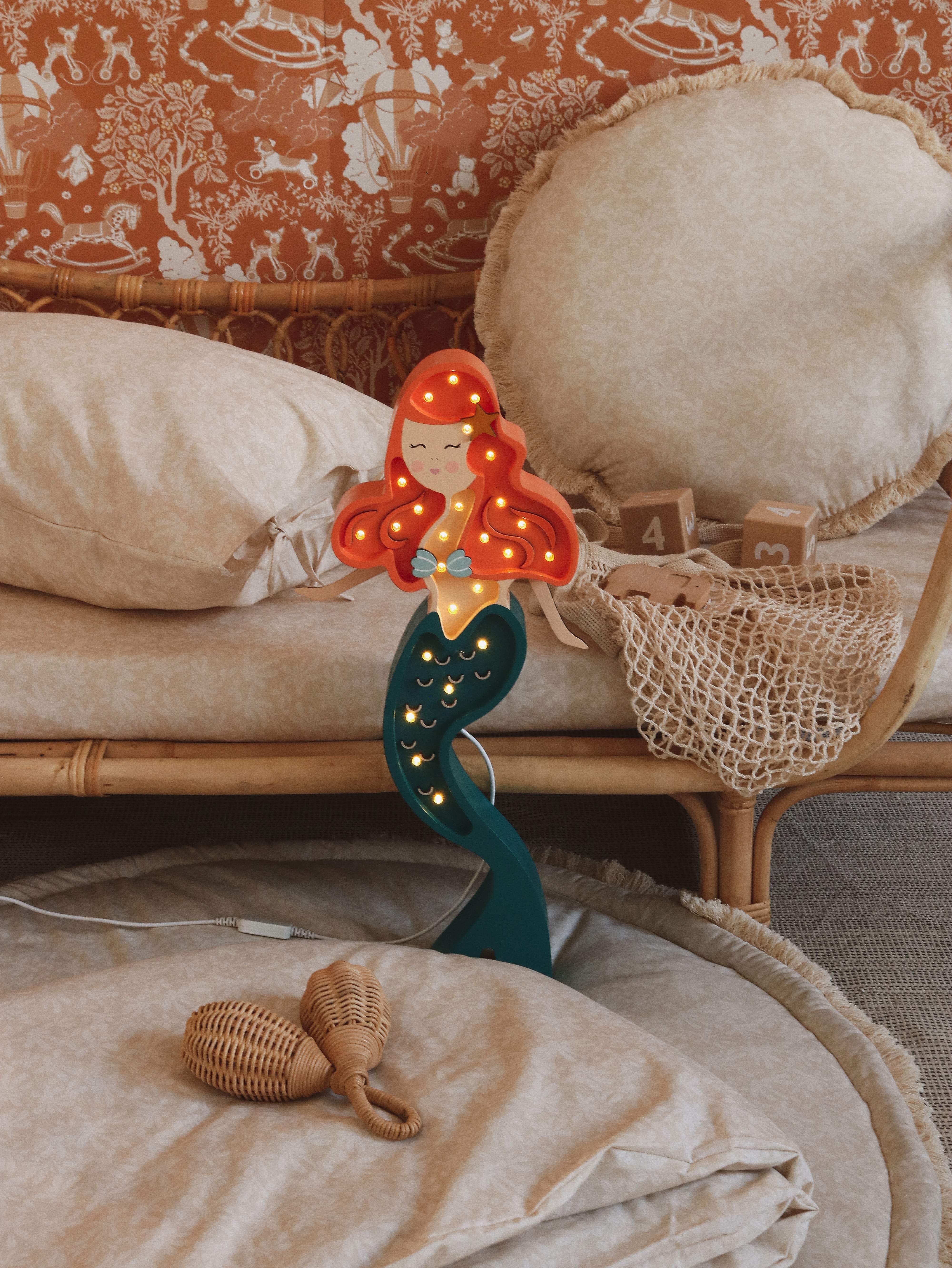 Eine skurrile Little Lights Ariel Mermaid Lampe mit roten Haaren und grünem Schwanz, die auf einem Nachttisch aus Bambus mit flauschigen Kissen und Rattan-Accessoires in einem gemütlich eingerichteten Zimmer steht.
