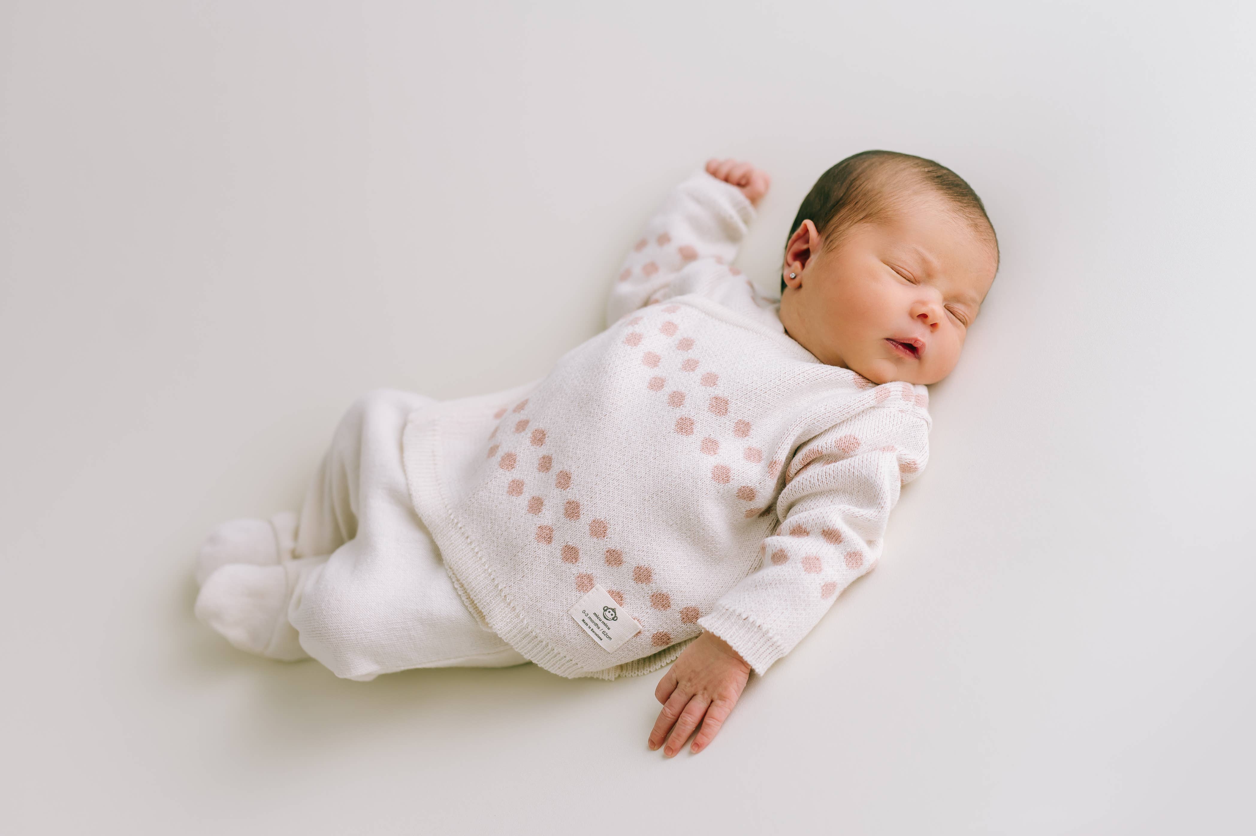 Ein schlafendes Baby, bekleidet mit einem Newborn Pack Punkte rosa: 0 -3 Monat (56 cm) von micu micu aus antiallergischer Baumwolle mit rosa Punkten, liegt auf einer weißen Oberfläche. Die Arme des Babys sind leicht angewinkelt und seine Augen sind geschlossen, ein friedlicher Ausdruck. Das Baby trägt Söckchen und liegt sanft auf dem Rücken.