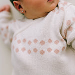 Nahaufnahme eines Babys, das einen beigen Strickpullover von Micu Micu aus dem Newborn Pack Punkte rosa: 0 -3 Monat (56 cm) trägt und rosa geometrische Formen um den Brustbereich aufweist. Das Baby hat kleine Ohrringe und liegt, wobei ein Teil seines Gesichts und Oberkörpers sichtbar ist.